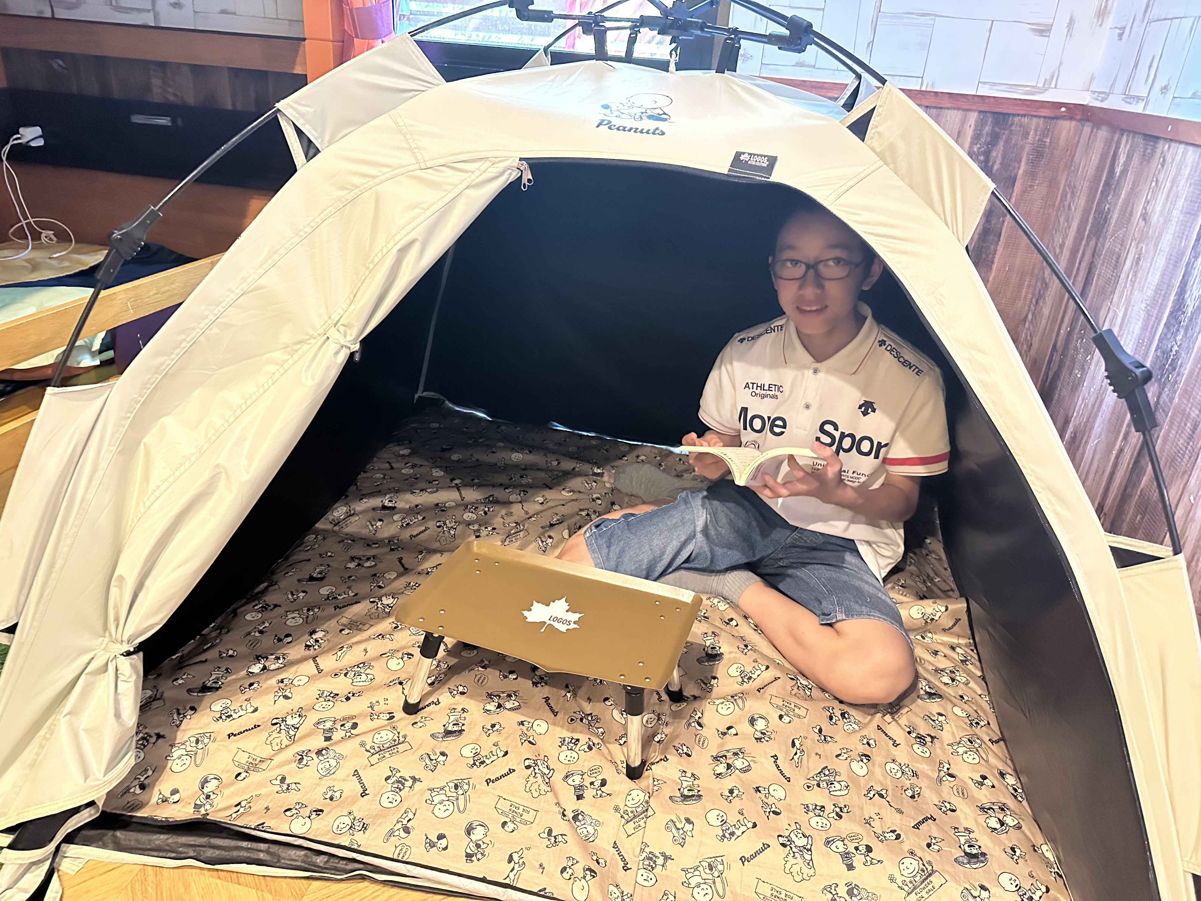 室内キャンプ風の宿泊室で、テントの中に座って本を開く生徒