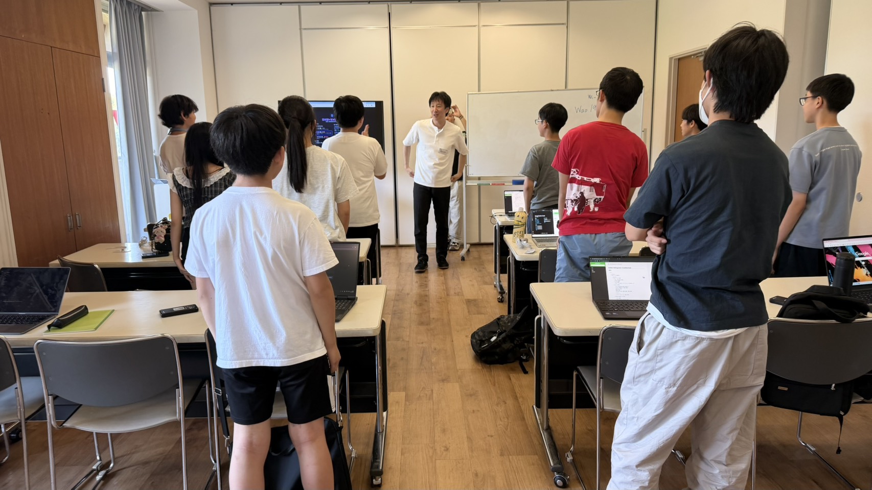 ノートPCが並ぶ研修室で立ち上がり、講師の話を聞く生徒たち