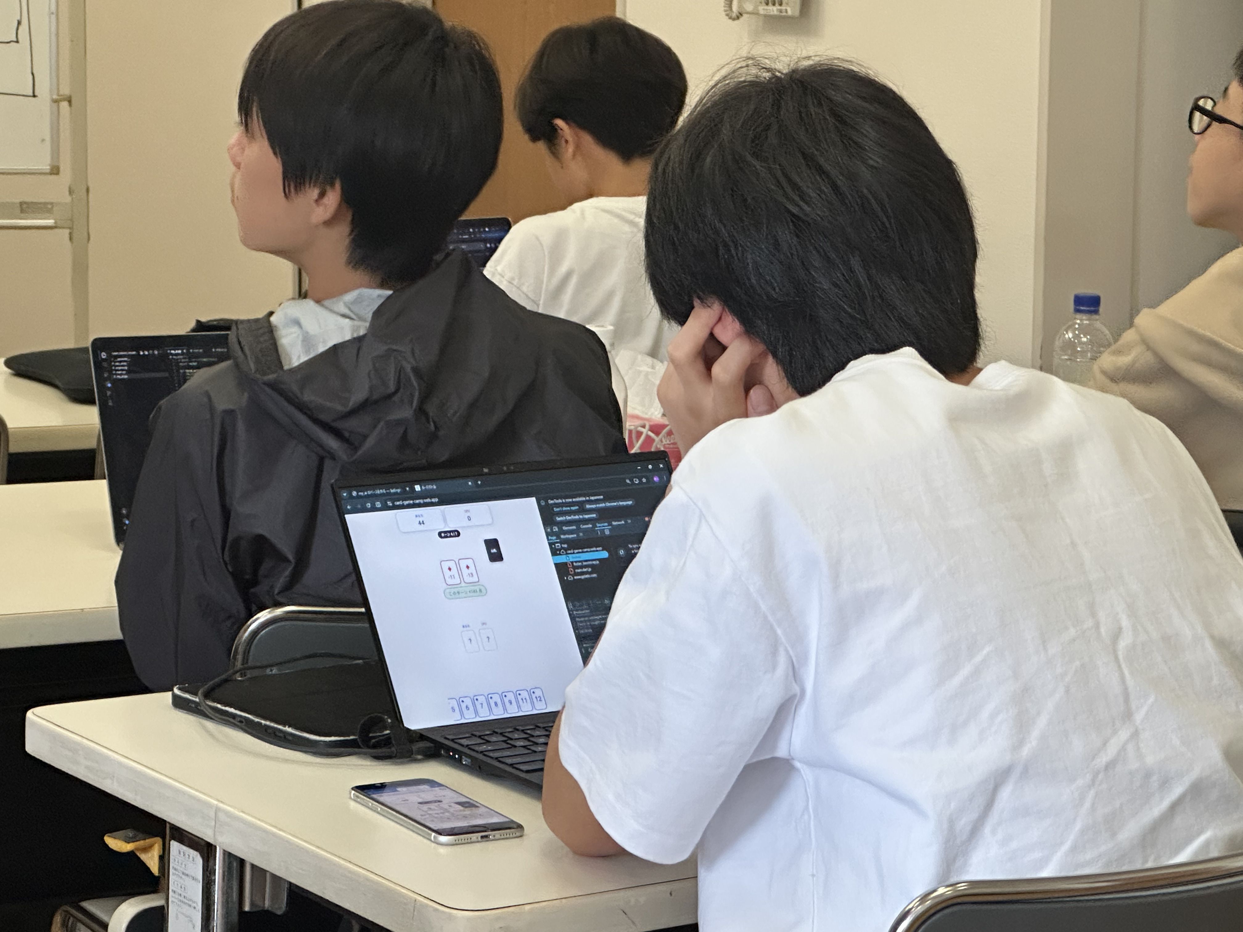 ノートPCでカードゲームの対戦画面を確認しながら個人開発に取り組む生徒