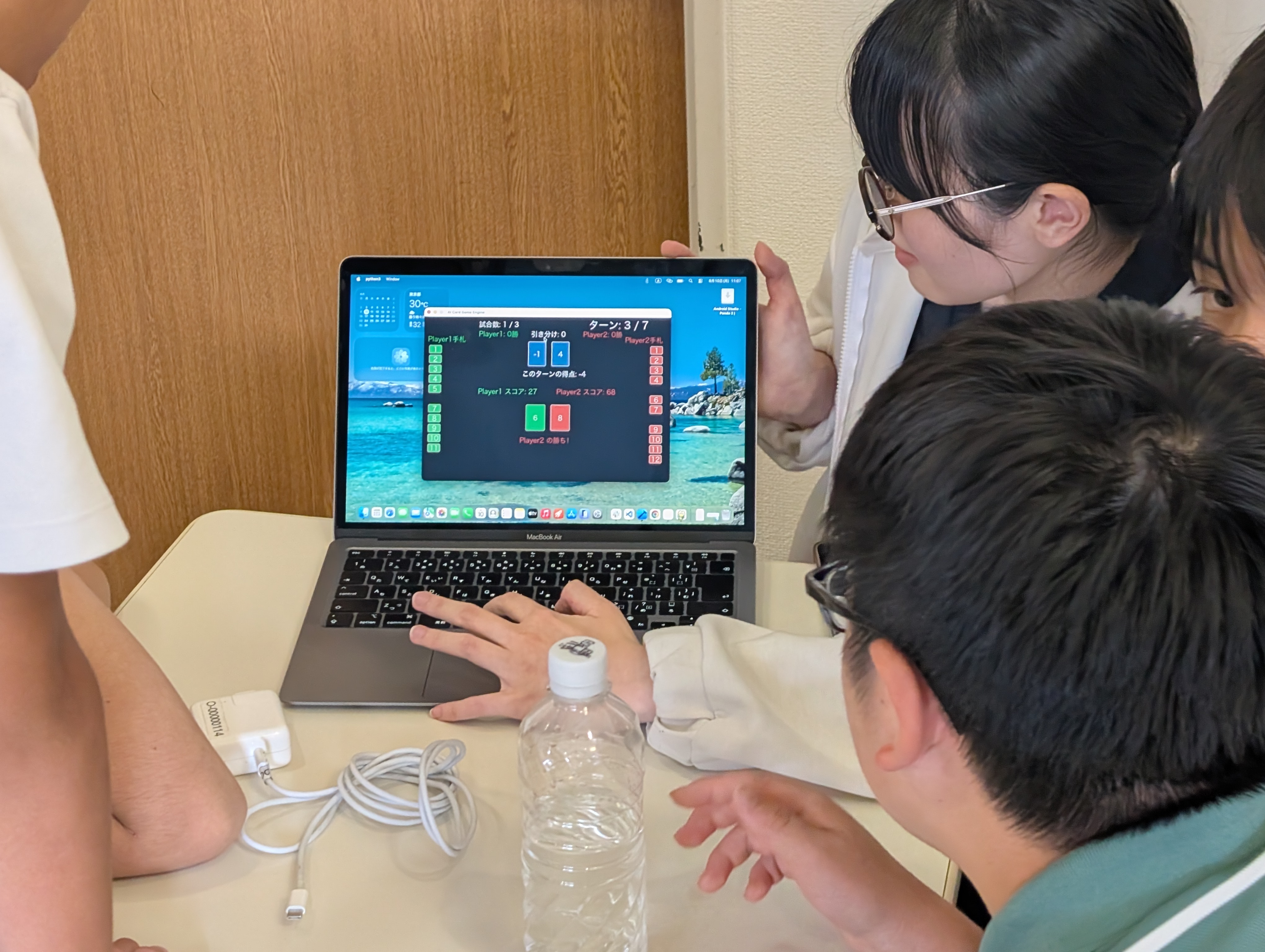 ノートPCに映るAI同士の対戦画面とスコアを、身を乗り出して見守る生徒たち